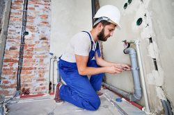 A man wearing a hard hat and blue overalls kneels on the floor, using a wrench to work on exposed plumbing pipes in a partially finished room—typical of skilled plumbers in Knoxville at work.